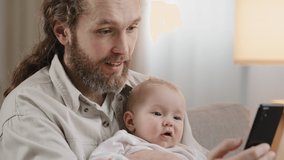 Bearded parent father daddy with little daughter son girl boy child baby kid infant newborn take photo on smartphone selfie picture with phone video chat online chatting in mobile app multitasking man