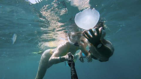 Slow motion young couple snorkeling underwater in the Red Sea, taking a cool selfie with a jellyfish. People dive into stunning tropical waters and take selfies
