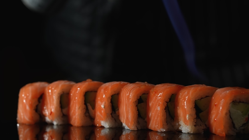 Sushi master roasts Philadelphia roll using burner. Sushi roll grill with salmon, avocado and cheese. Fire burning salmon close up. Cooking traditional Japanese food with seafood and fish. Slow motion