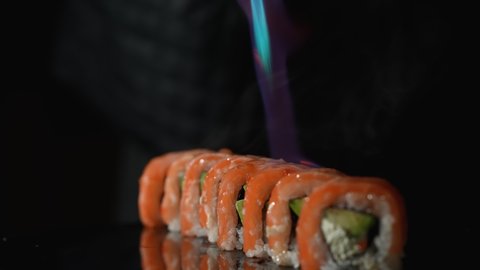 Sushi master roasts Philadelphia roll using burner. Sushi roll grill with salmon, avocado and cheese. Fire burning salmon close up. Cooking traditional Japanese food with seafood and fish. Slow motion