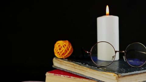 Burning candle with eyeglasses standing on old books. Big white candle burning on black background surrounded by decor. Concept of reading in dark room in illumination of flame.
