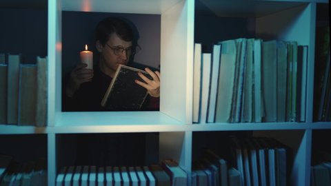 Man in glasses with candle reading book, standing behind bookshelf. Male holds burning candle while reading book in wooden bookcase with various literature in dark room.