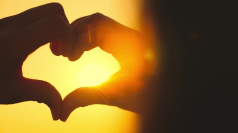 Hands of woman pose making romantic heart sign. Female person shows love symbol with fingers. Heart as shadow pantomime against sky with orange sunset extreme closeup