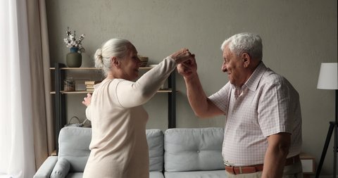 Carefree healthy older 75s spouses holds hands dance in living room. Active senior wife husband couple in love enjoy hobby listen music at home. Untroubled retired life, relationships, harmony concept