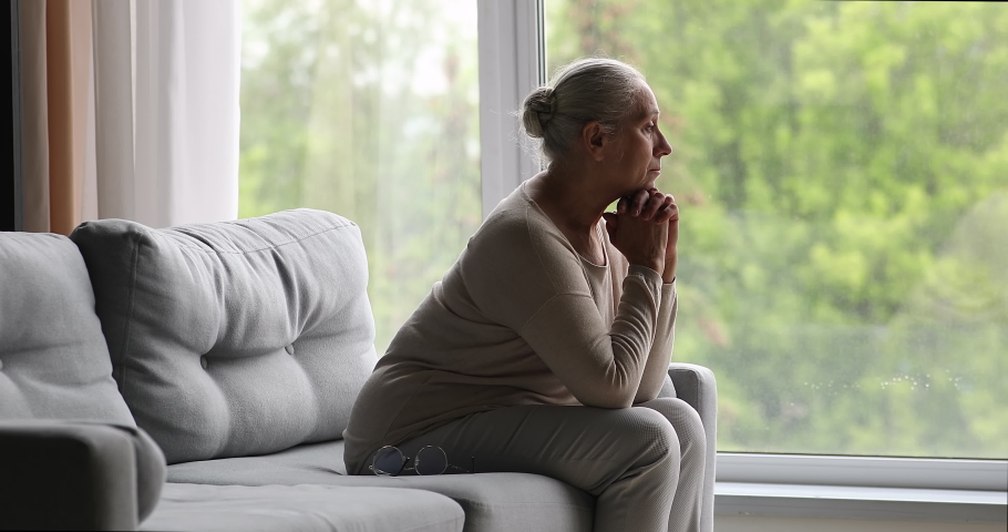 Side view 65s widow lonely grey-haired woman sits on sofa in living room looking into distance, feeling solitude, recollects past, missing longing for husband. Retiree in nursing home, ageing concept Royalty-Free Stock Footage #1087300037