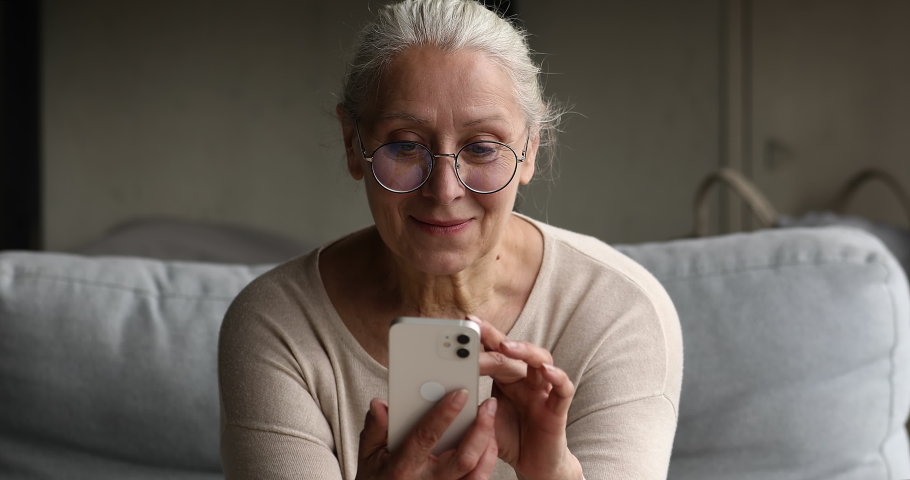 Elderly 65s woman sit on couch in living room with cell phone, wear glasses looks at smartphone screen, web surfing information enjoy easy modern tech usage spend free time at home. Connection concept Royalty-Free Stock Footage #1087300139