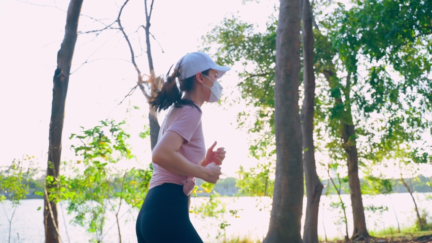 Asian young active sport woman wear mask run on street in public park. Beautiful athlete girl in sportswear exercise by jogging workout outdoor for health during pandemic in evening sunset in garden.