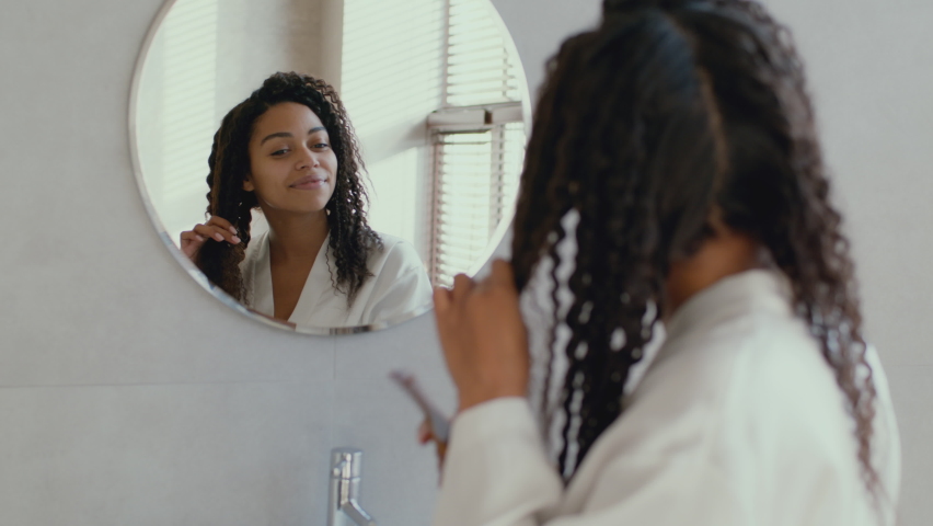 Domestic hairstyling. Over shoulder portrait of young peaceful curly african american lady wearing bathrobe combing her hair, looking at mirror at bathroom in morning, zoom in, slow motion Royalty-Free Stock Footage #1087310591