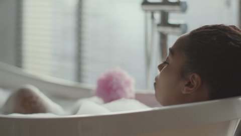 Relax and hygiene concept. Young african american woman taking foamy bath, washing her leg with sponge, over shoulder view, tracking shot, slow motion, free space