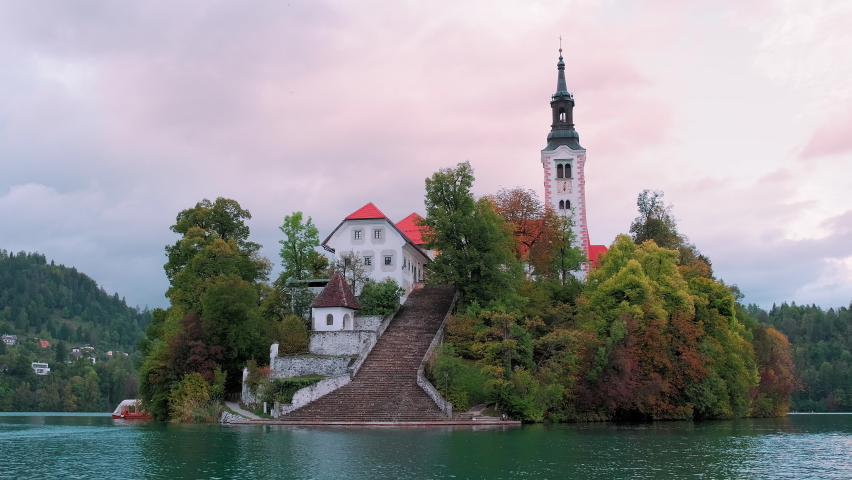 Lake Bled in Slovenia from boat. Sunset view on Assumption of Maria Church against cloudy Julian Alpine mountains. Church reflecting in lake Bled waters. Handheld shot rotating around church. | Shutterstock HD Video #1087312526
