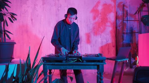 Modern DJ of Asian Appearance Performs a Music Track in Neon Lighting. Working at the Mixer, a Man Puts on Records, Sound Tracks, Individual Sounds and Makes a Mix Out of Them. 