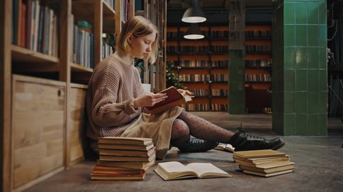 Booklover concept. Young serious lady student reading book, sitting among stack of books on library floor, side view, empty space, slow motion