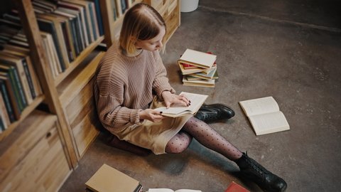 Bookworm concept. Top view of young focused woman student sitting on library floor with stack of books and reading, tracking shot, slow motion