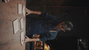 Vertical video of confident young craftsman stands near a work table in a carpentry workshop. A man in a working apron shakes his hands from chips and crosses his arms over his chest, general plan.