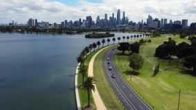 Beautiful view of Melbourne CBD city skyline from Albert Park Lake, aerial drone view with slow cinematic pan. View of the lake, race track road with cars, people exercising, walking track and park.