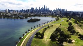 Beautiful view of MELBOURNE CITY skyline from ALBERT PARK LAKE. Drone video of lake, park, walking path, road, palm trees and the Melbourne CBD or downtown district on a cloudy day, mid day sun.