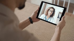 Over shoulder view of Indian man holding digital tablet and having online videocall with virtual doctor while staying sick at home. Close up of patient in video conferencing with general practitioner.