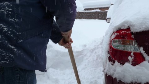 A man in a cap, jacket and jeans is shoveling snow off the area near his home. Nearby is a red car covered with snow during a snow storm. Snowfall in the background