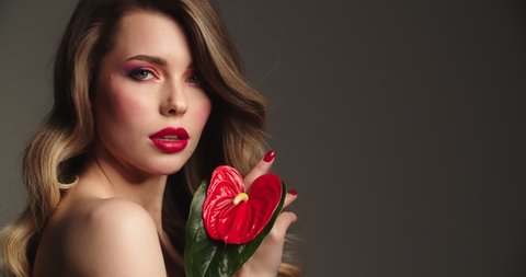 Fashion model with  hair blowing in the wind. Sexy blonde woman with red lips holds a red flower. Closeup face of a pretty young woman with bright makeup.  Slow motion.