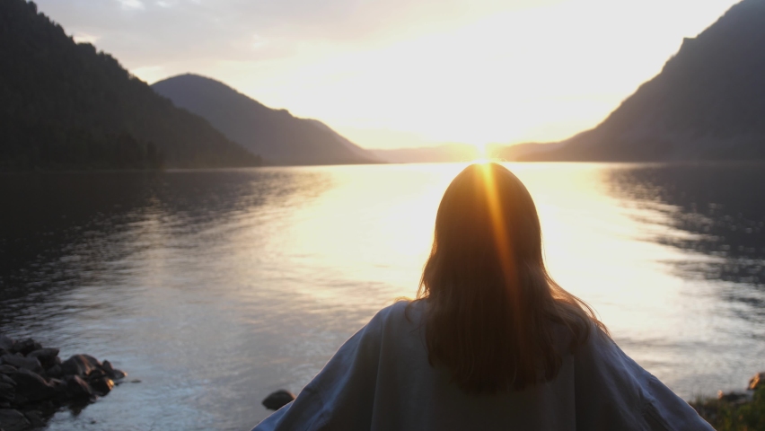Woman raising hands and makes heart on the edge of the mountain cliff on lake at sunset