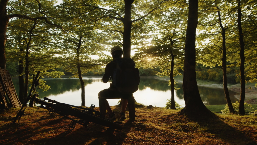 boy with backpack sits near the lake at sunset
