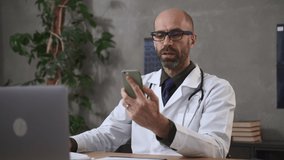 Male doctor having video call with phone in hand and sitting at table in modern clinic spbas. Bearded middle aged man holds smartphone in hand and looks at screen, speaks with smile and sits at desk