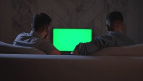 Two male fans watching football and sitting on sofa in apartment interior spbas. 4k video Back view of young american men watch tv match in front of screen and talk with smiles, cheer for team and sit