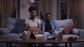 Young couple watching match and cheering for sports team while sitting on couch at home spbas. 4k video African American people watch tv and have fun, wave and sit on sofa in living room, food on