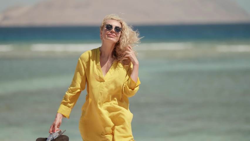 a blonde in sunglasses and a yellow fluttering dress holds hair and walks against the background of a blurred sea