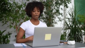 Confident young African-American woman talking and gesturing during online remote video conference call sitting at desk in light home office room with modern biophilic interior design, slow motion.