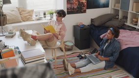 High-angle of Caucasian guy wearing hoodie, sitting with legs up on desk in his room in afternoon, holding book, turning around and talking to Black female school friend who using computer, laughing - Powered by Shutterstock - Get 15% off with code: PIKWIZARD15