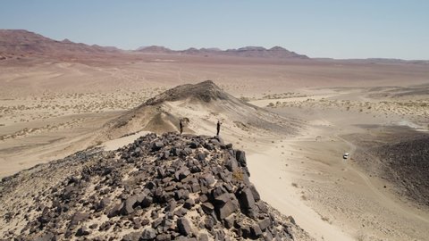 Two guys atop rocky outcrop next to high dune in Namibian wilderness; aerial
