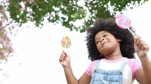 Little girl eating lollipops sweet rainbow candy in the park with excited mood. Delicious snack with sugar. Concept of toy and learning for children