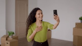 Cheerful Lady Video Calling Moving New Home Standing Among Moving Boxes And Waving Hello To Smartphone Indoor. Relocation, Real Estate Ownership, Modern Distance Communication Concept