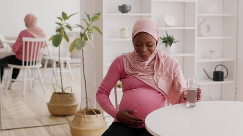 Pregnant African American Woman Drinking Water From Glass Hydrating Caring For Health During Pregnancy Sitting Wearing Headscarf At Home. Healthy Hydration, Childbirth And Healthcare. Slow Motion