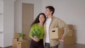 Real Estate. Joyful Couple Holding Moving Box And Green Plant In Pot Celebrating Relocation Hugging Standing In New House Indoor Among Cardboard Boxes. Apartment Rental, Purchase And Ownership