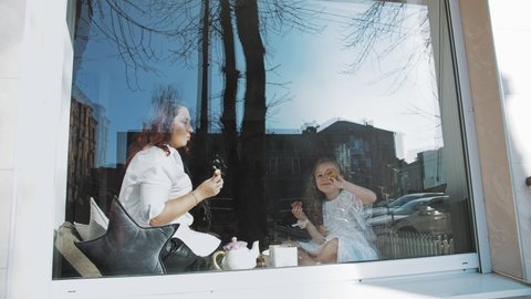 tea party. mother and little daughter are drinking tea with sweets, cookies, sitting by the window, on a large windowsill. view through the window, from outdoors