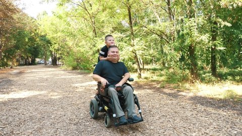 father's day. person with a disability. dad and his little cute son are walking in park, on summer day. dad is a wheelchair user. people with special needs.