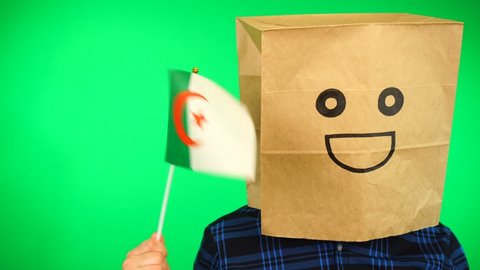 Portrait of man with paper bag on head waving Algerian flag with smiling face against green background.