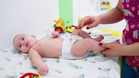 KYIV, UKRAINE - August 2021: Baby boy is at surgeon’s check-up. Female doctor examining baby’s legs and knees. Toys at the backdrop.