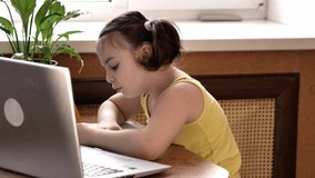 A little girl with two tails sits at a laptop, diligently writes in a notebook, pronounces the words. Online education for children remotely with a computer.