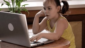 A little girl with two ponytails is sitting at a laptop, restless, her attention scattered. Distance learning for children, attention deficit disorder.