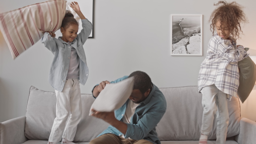 Slowmo shot of joyful African-American man and his two cute little daughters pillow fighting and having fun together at home