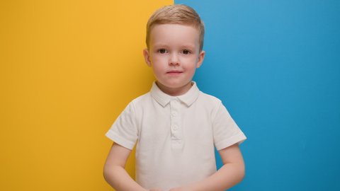 Small blond boy expresses love by showing heart sign with his hands near his chest while standing on blue-yellow studio background. Call to love, hands in shape of heart, Ukraine with love.