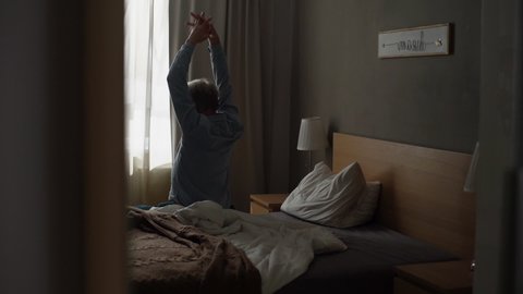 Remote back view of unrecognizable mature male warming up sitting on bed in morning after waking up at home. Senior adult man stretching hands up, massaging neck muscles sitting by window in morning.の動画素材