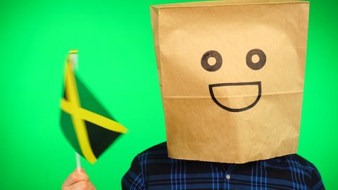 Portrait of man with paper bag on head waving Jamaican flag with smiling face against green background.