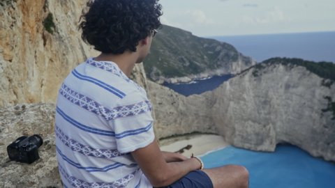 Young man admires Navagio Shipwreck Cove Greece from cliffs above, slow motion