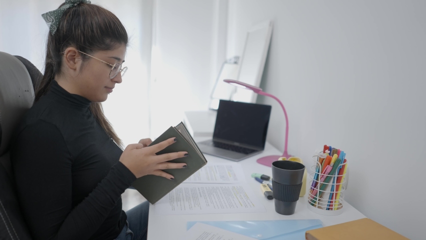 A young student of Colombian universitary sitting at table reading a book and writes notes in her paper.