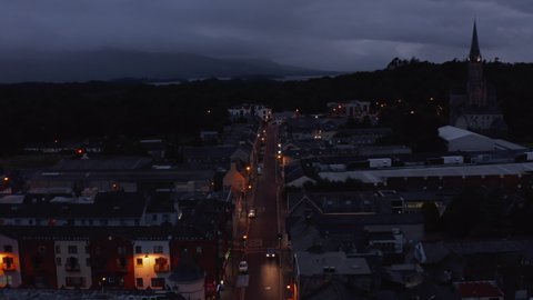 Backwards reveal of straight illuminated street in evening town. Overcast sky and lake in distance. Killarney, Ireland