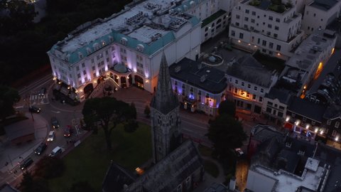 High angle view of old medieval church tower in evening town. Tilt up reveal of arranged park garden and hazy hills in distance. Killarney, Ireland